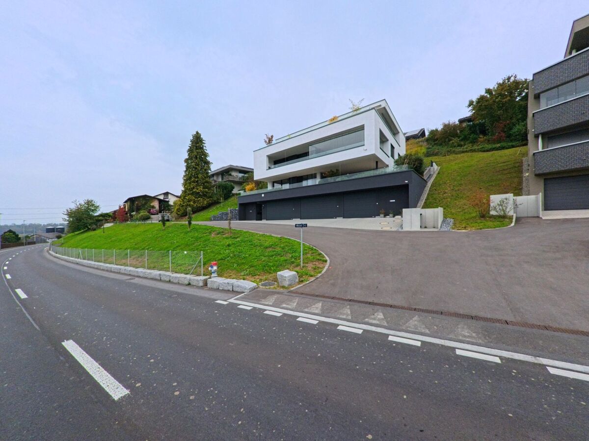 An einer ansteigenden Zufahrt in Hueenenberg (6331, Kanton Zug) steht ein modernes Hanghaus mit grossen Terrassen, waehrend die Strasse in einer Kurve vorbeifuehrt.