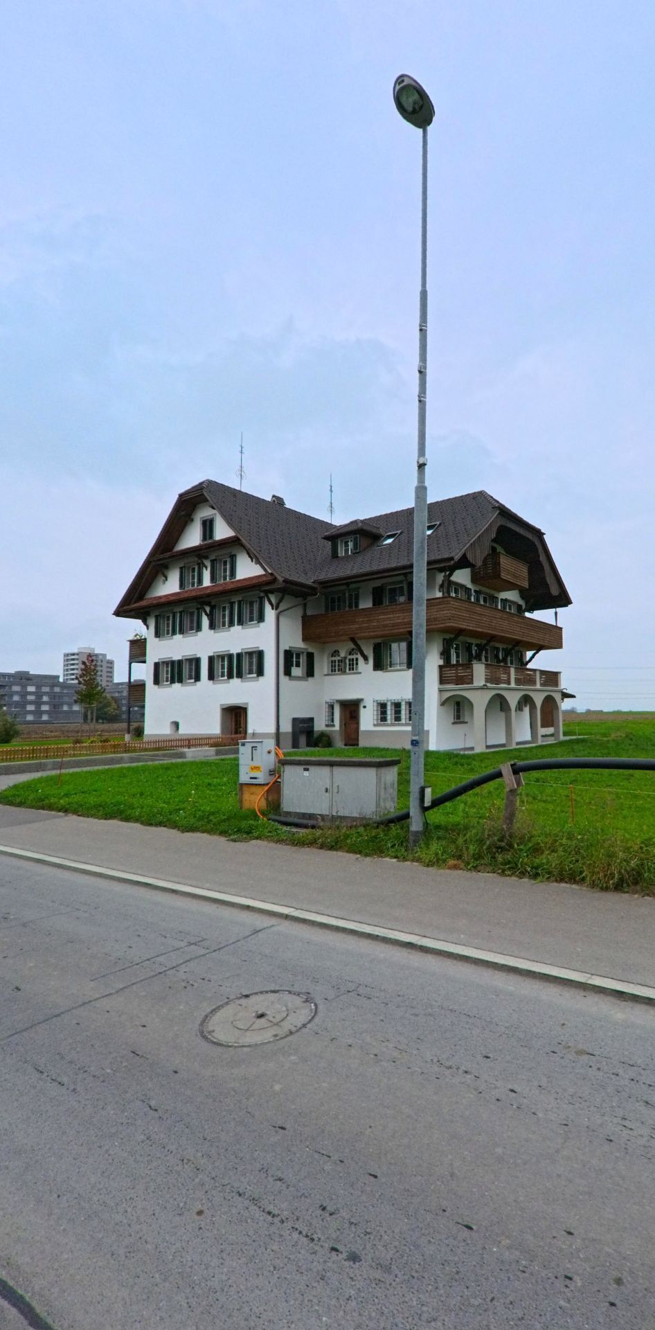 Am Wiesenrand von Hueenenberg (6331, Kanton Zug) steht ein grosses traditionsreiches Haus mit dunklem Satteldach und Holzbalkonen neben einer Strassenlaterne.