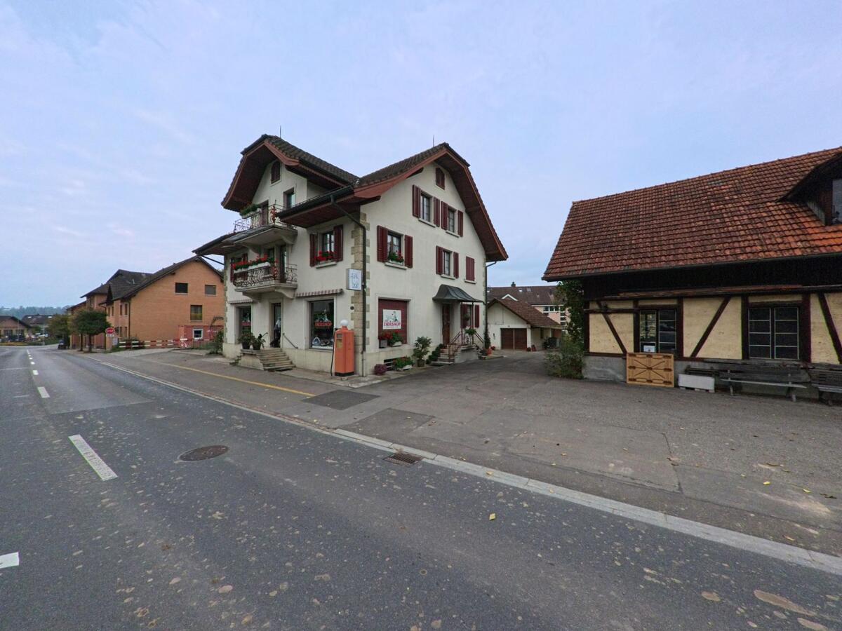 Ein traditionsreiches Wohnhaus mit roten Fensterlaeden steht in Hueenenberg (6331, Kanton Zug) direkt an der Hauptstrasse neben einem aelteren Anbau.