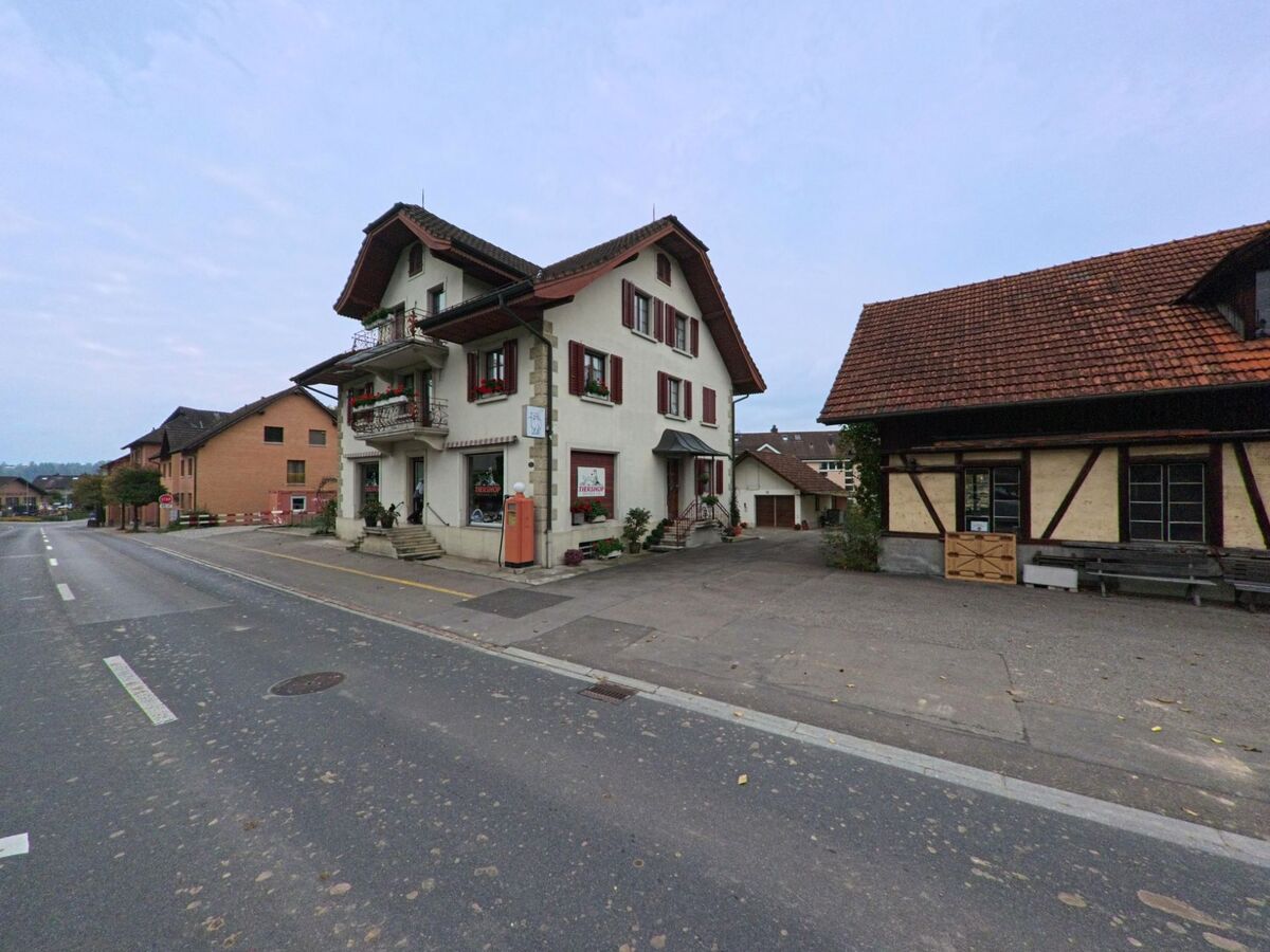 Ein traditionsreiches Wohnhaus mit roten Fensterlaeden steht in Hueenenberg (6331, Kanton Zug) direkt an der Hauptstrasse neben einem aelteren Anbau.