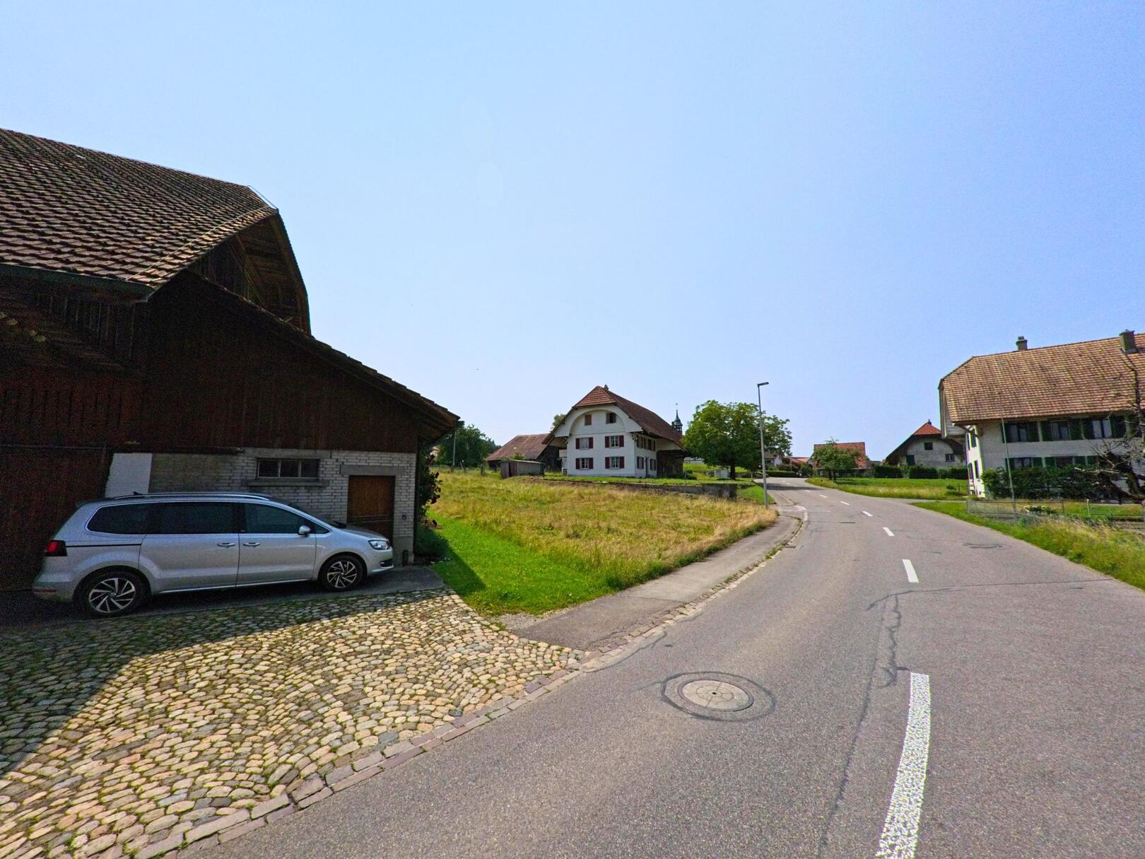 In Hüniken (4554, Kanton Solothurn) verläuft eine Strasse an traditionellen Häusern entlang, mit einem geparkten Auto und einer grünen Wiese im Vordergrund.