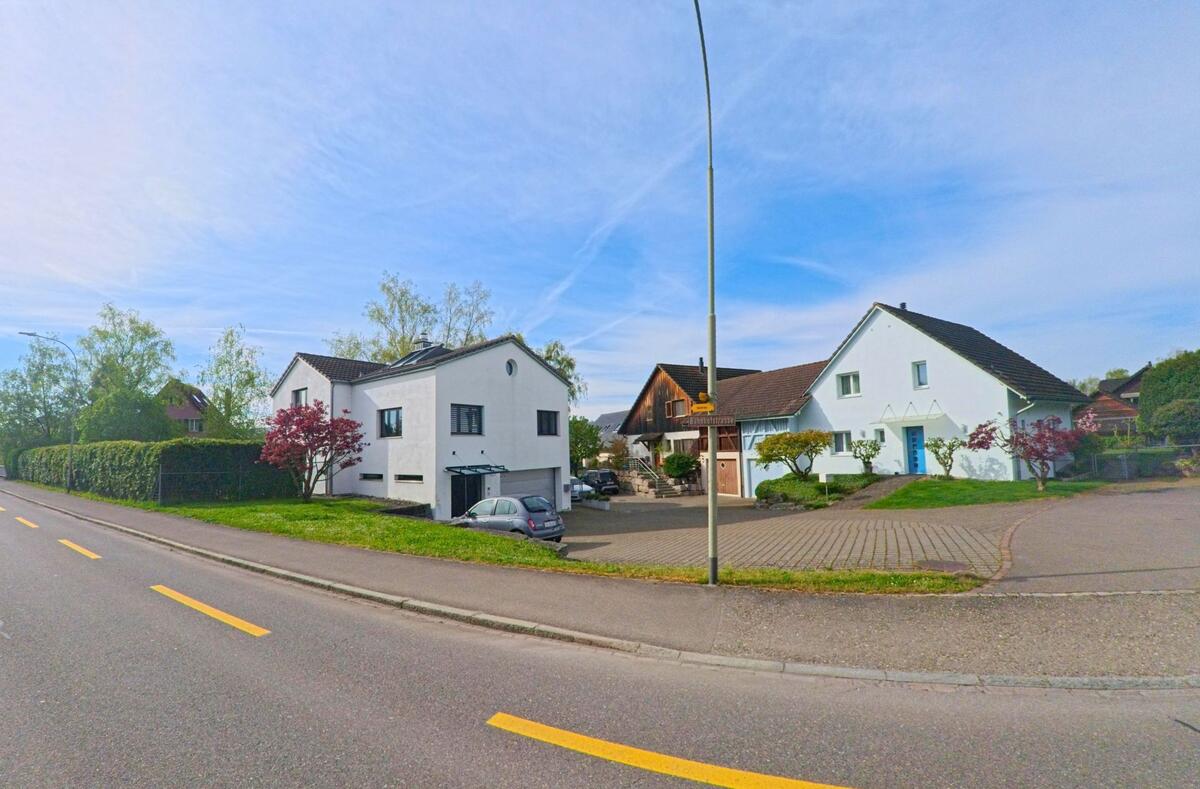 In Hüntwangen im Kanton Zürich mit der PLZ 8194 stehen mehrere freistehende Einfamilienhäuser mit gepflegten Vorgärten entlang einer gut ausgebauten Strasse mit gelber Fahrbahnmarkierung.