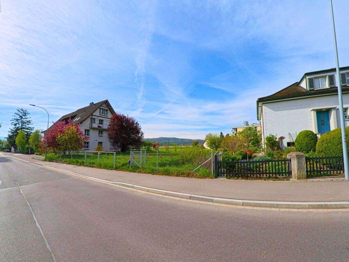 Entlang einer ruhigen Strasse in Hüntwangen im Kanton Zürich mit der PLZ 8194 stehen Wohnhäuser mit gepflegten Gärten, im Hintergrund öffnet sich der Blick auf Felder und bewaldete Hügel.