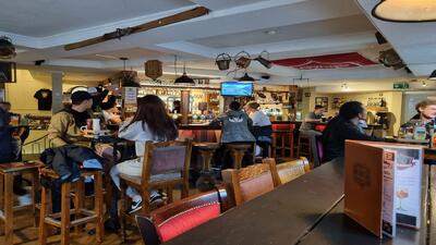 Im Hueesi Bierhaus in Interlaken (3800, Kanton Bern) sitzen Gaeste an der gemuetlichen Bar mit Holzmoebeln und verfolgen eine Fernsehsendung in geselliger Atmosphaere.