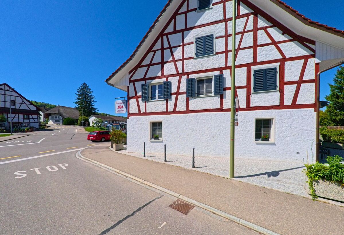 Im Zentrum von Hüttikon im PLZ-Gebiet 8115 im Kanton Zürich steht ein weisses Fachwerkhaus mit roten Balken und dem Restaurant „Alte Post“ an einer Kreuzung mit STOP-Markierung.