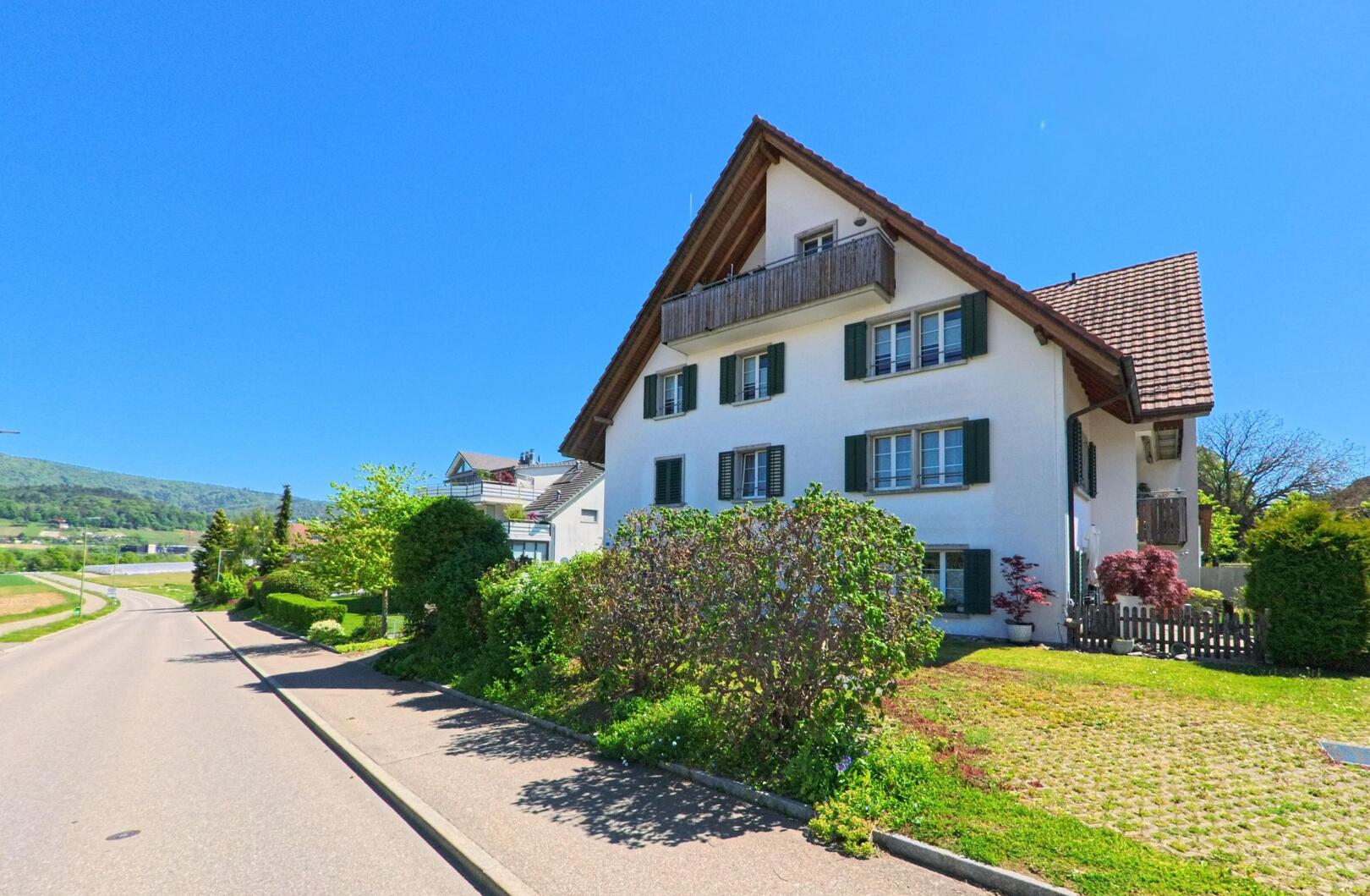 Ein traditionelles Wohnhaus mit grünem Fensterläden steht in Hüttikon im PLZ-Gebiet 8115 im Kanton Zürich an einer ruhigen, sonnigen Strasse mit Blick auf das Umland.