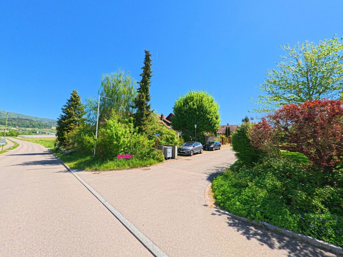 In Hüttikon im PLZ-Gebiet 8115 im Kanton Zürich zweigt eine ruhige Wohnstrasse mit parkenden Autos und gepflegtem Grün von der Hauptstrasse ab.