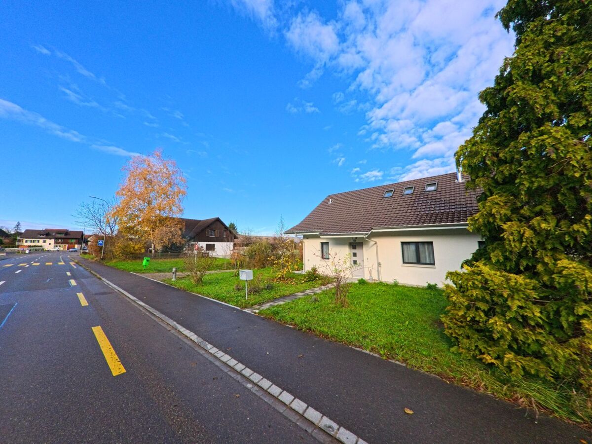 Am Rand einer Wohnstrasse in Hüttlingen (8553, Kanton Thurgau) steht ein flaches Einfamilienhaus mit Rasenfläche und Blick auf herbstliche Bäume.