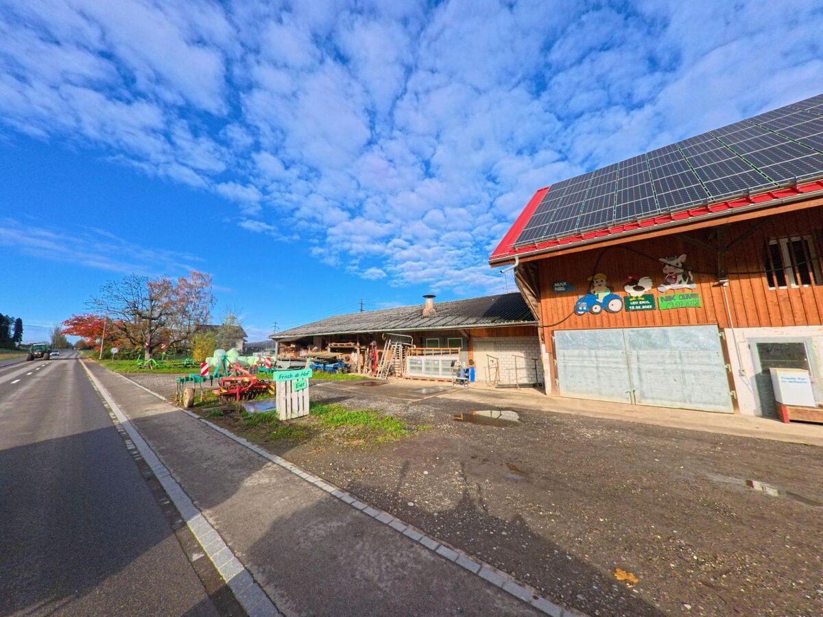 An einer Landstrasse in Hüttlingen (8553, Kanton Thurgau) steht ein Hofgebäude mit Solarpanels, davor sind landwirtschaftliche Geräte und ein kleiner Verkaufsstand aufgebaut.