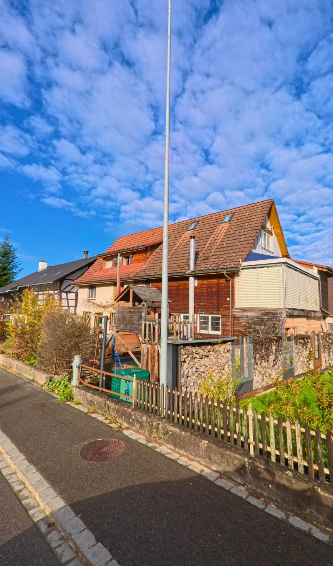 Hinter einem niedrigen Gartenzaun in Hüttlingen (8553, Kanton Thurgau) steht ein Holzhaus mit Ziegeldach und gestapeltem Brennholz am Rand des Gehwegs.