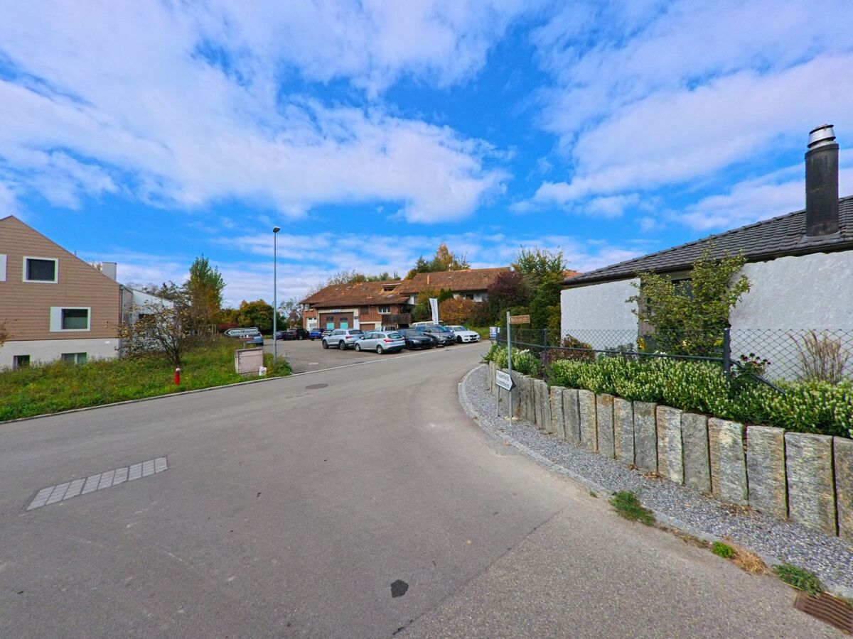 Eine ruhige Quartierstrasse führt in Hüttwilen (8537, Kanton Thurgau) an einem Parkplatz und gepflegten Vorgärten unter blauem Himmel vorbei.