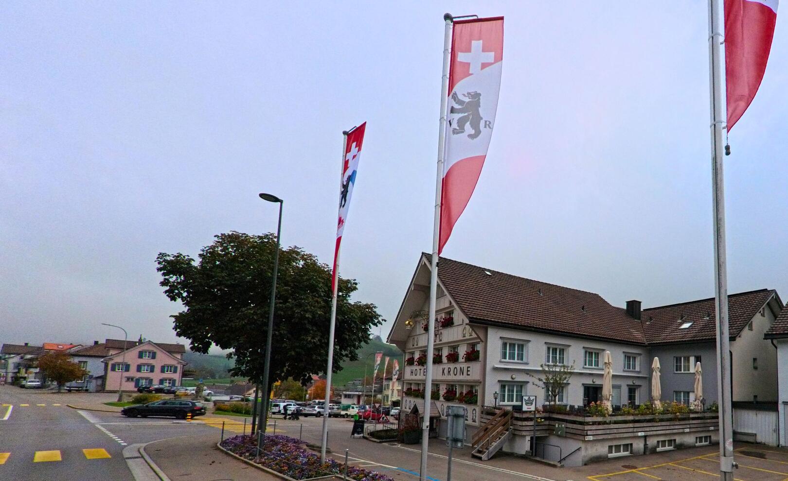 Vor dem Hotelgebäude in Hundwil (9064, Kanton Appenzell Ausserrhoden) stehen mehrere Fahnen, waehrend im Hintergrund Wohnhaeuser und ein leicht bewoelkter Himmel zu sehen sind.
