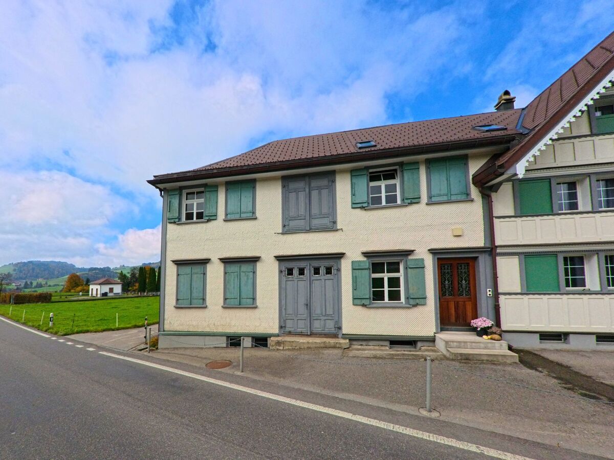 Ein Wohnhaus mit grünen Fensterläden befindet sich an einer schmalen Dorfstrasse in Hundwil (9064, Kanton Appenzell Ausserrhoden).
