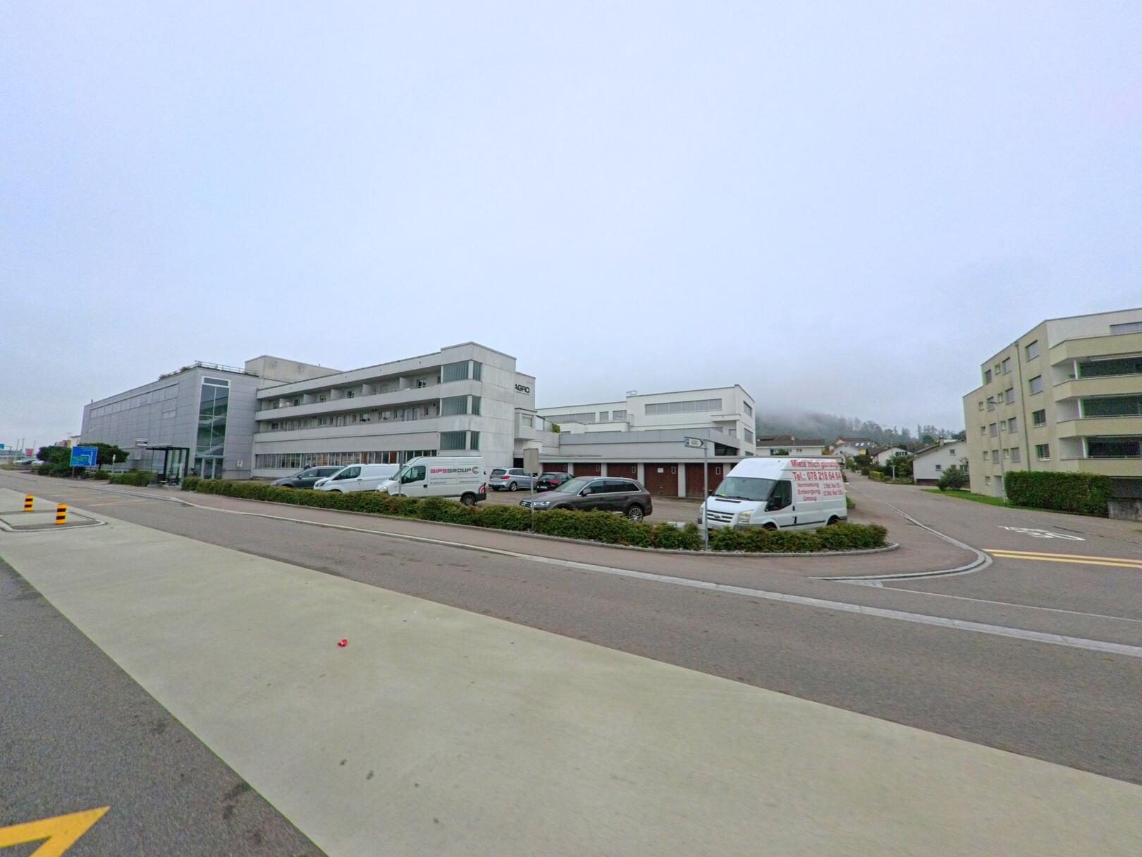 In Hunzenschwil (5502, Kanton Aargau) stehen moderne Gewerbe- und Wohnbauten entlang einer breiten Strasse mit geparkten Fahrzeugen.
