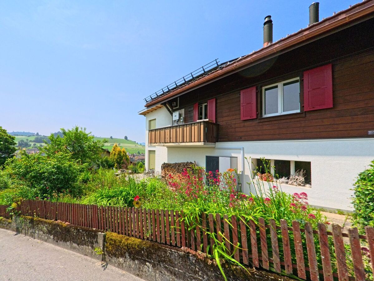 In Huttwil (4950, Kanton Bern) ist ein gepflegtes Haus mit einem bunten Garten und einem Zaun entlang der Straße zu sehen, vor einer sanften Hügelkulisse.
