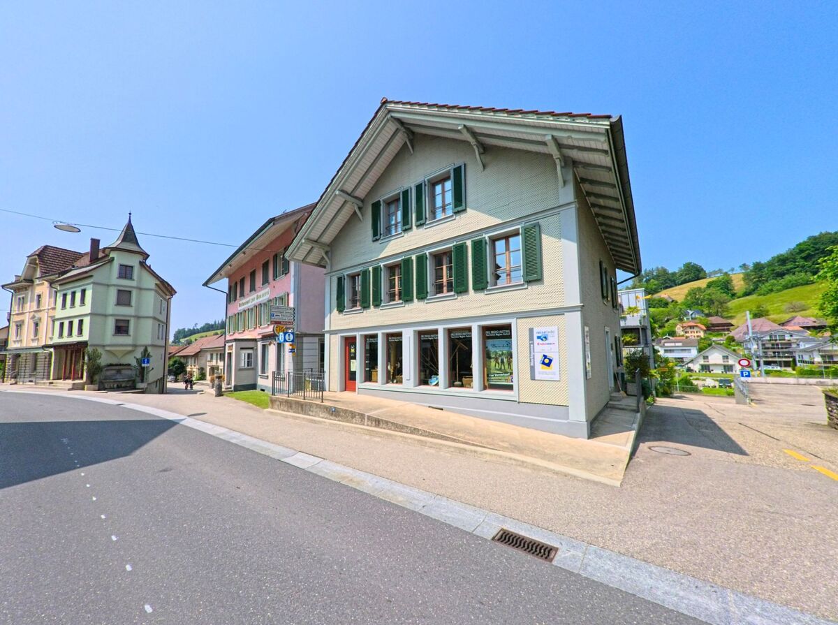 In Huttwil (4950, Kanton Bern) ist ein historisches Gebäude mit grünen Fensterläden an einer breiten Straße zu sehen, während die umliegenden Hügel im Hintergrund sichtbar sind.