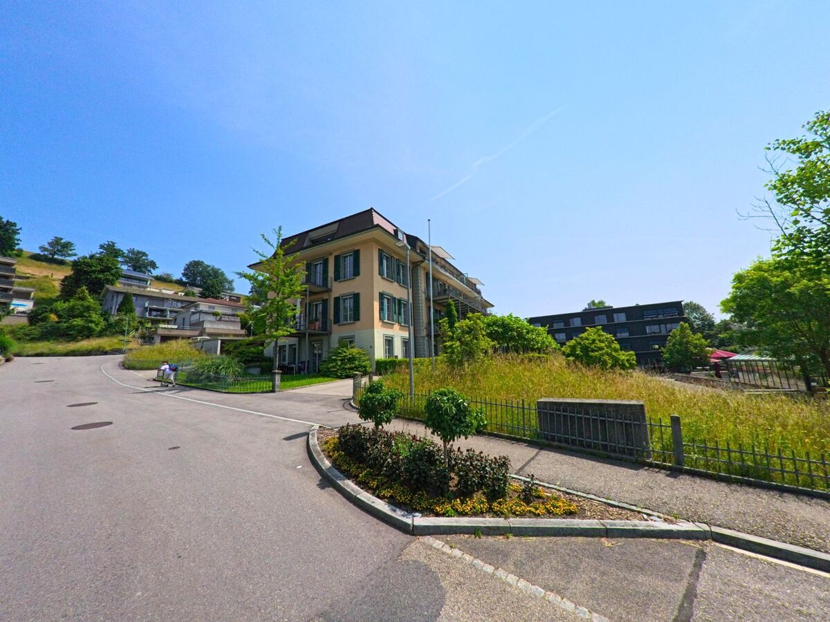 In Huttwil (4950, Kanton Bern) führt eine steile Straße an einem klassischen Mehrfamilienhaus vorbei, während moderne Gebäude im Hintergrund sichtbar sind und grünes Gras die Szene ergänzt.