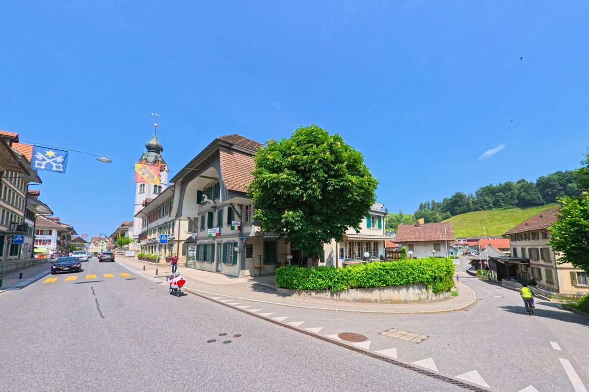 In Huttwil (4950, Kanton Bern) ist eine belebte Straße zu sehen, die an traditionellen Häusern entlangführt, mit einem Blick auf den Kirchturm und die grünen Hügel im Hintergrund.