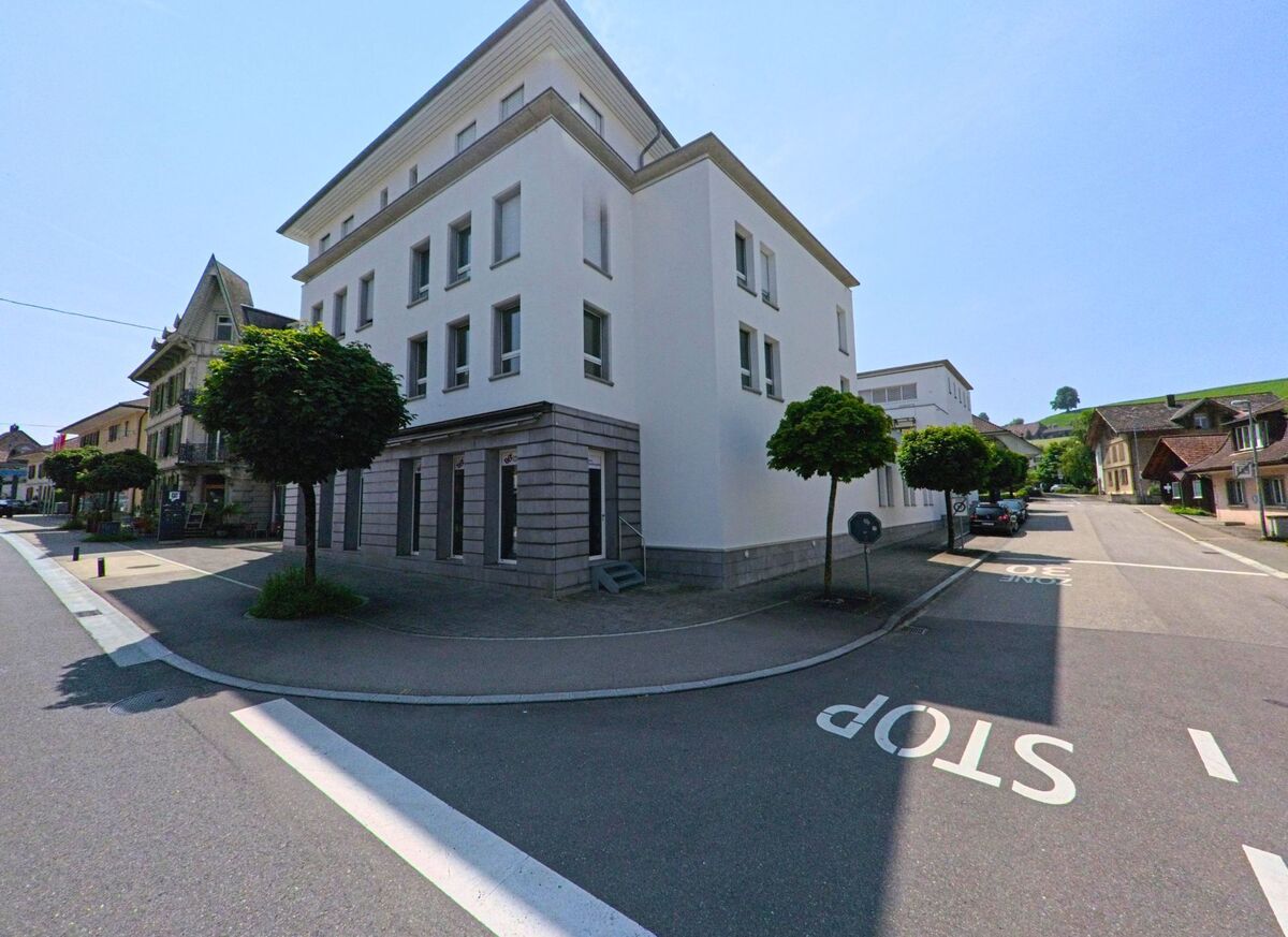 In Huttwil (4950, Kanton Bern) sind moderne und traditionelle Gebäude entlang einer breiten Straße zu sehen, mit Bäumen und einer gut markierten Stopplinie im Vordergrund.