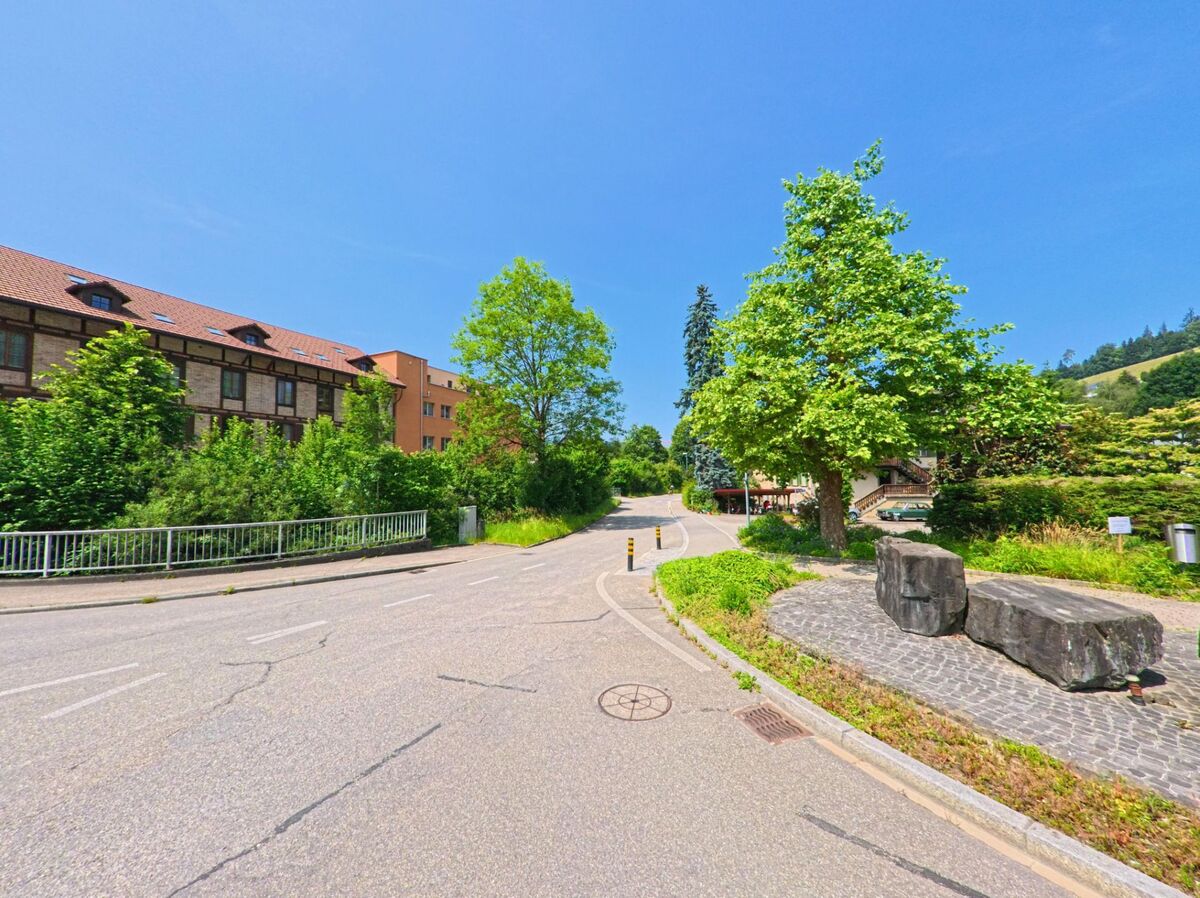 In Huttwil (4950, Kanton Bern) führt eine ruhige Straße mit grünen Bäumen und Sträuchern an einem modernen Gebäude entlang, während ein kleiner Steingarten an der Seite zu sehen ist.