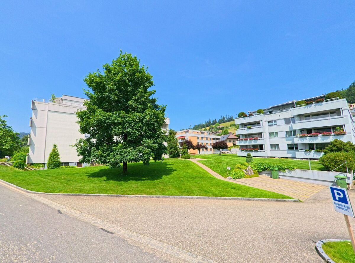 In Huttwil (4950, Kanton Bern) ist ein moderner Wohnblock mit gepflegtem Grünflächen und Bäumen entlang der Straße zu sehen, während die sanfte Hügelkette im Hintergrund sichtbar ist.