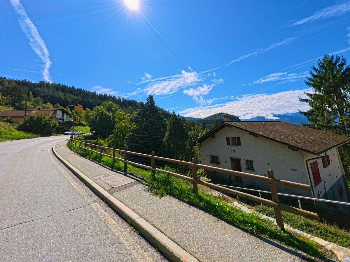 Eine Strasse mit Holzgeländer fuehrt in Icogne (1977, Kanton Wallis) an einem weissen Haus mit roten Fensterlaeden vorbei, umgeben von Wald und Bergen.