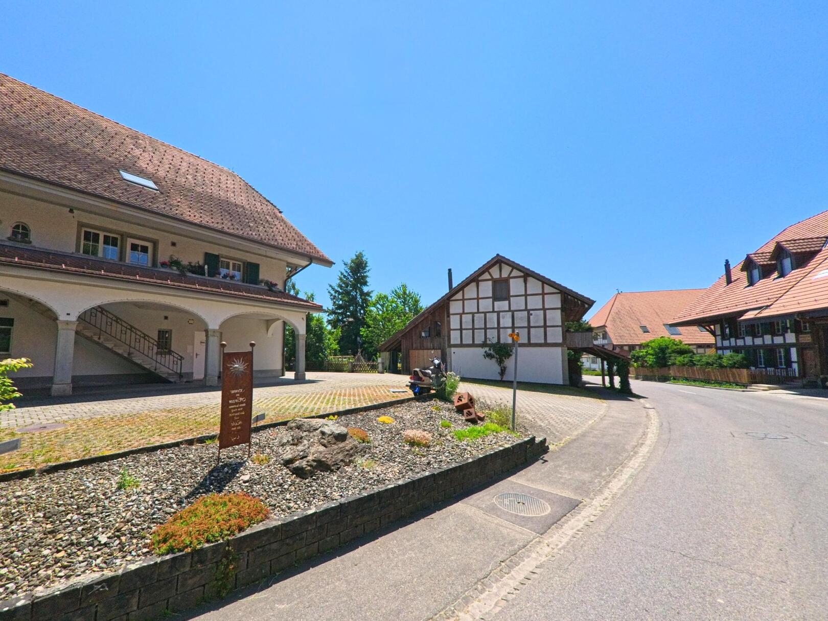 Ein traditionelles Fachwerkhaus und ein modernes Gebäude in Iffwil (3305, Kanton Bern) stehen an einer Strasse, die von einem schön angelegten Garten gesäumt ist.