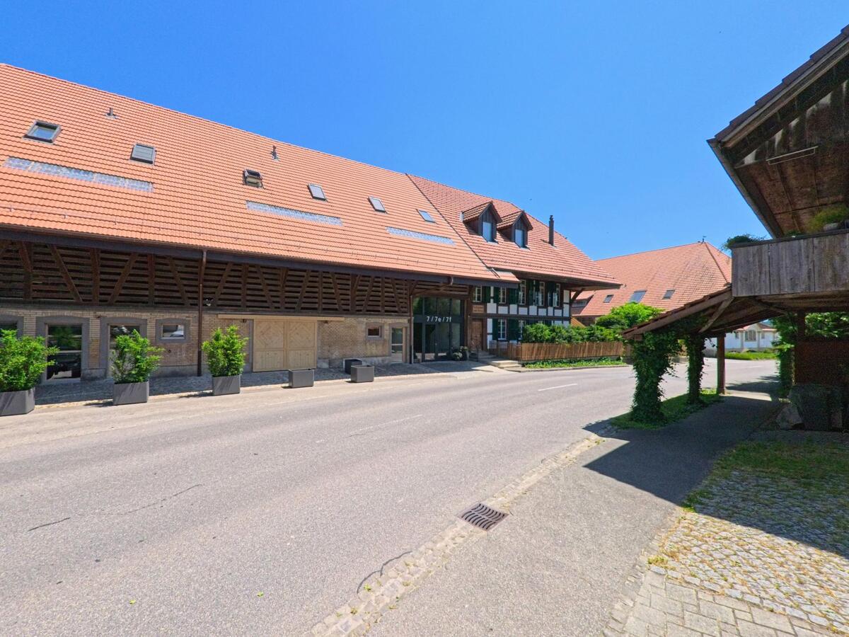 Ein modernes Gebäude mit einem steilen Dach in Iffwil (3305, Kanton Bern) steht an einer Straße, gesäumt von grünen Pflanzen in großen Töpfen.