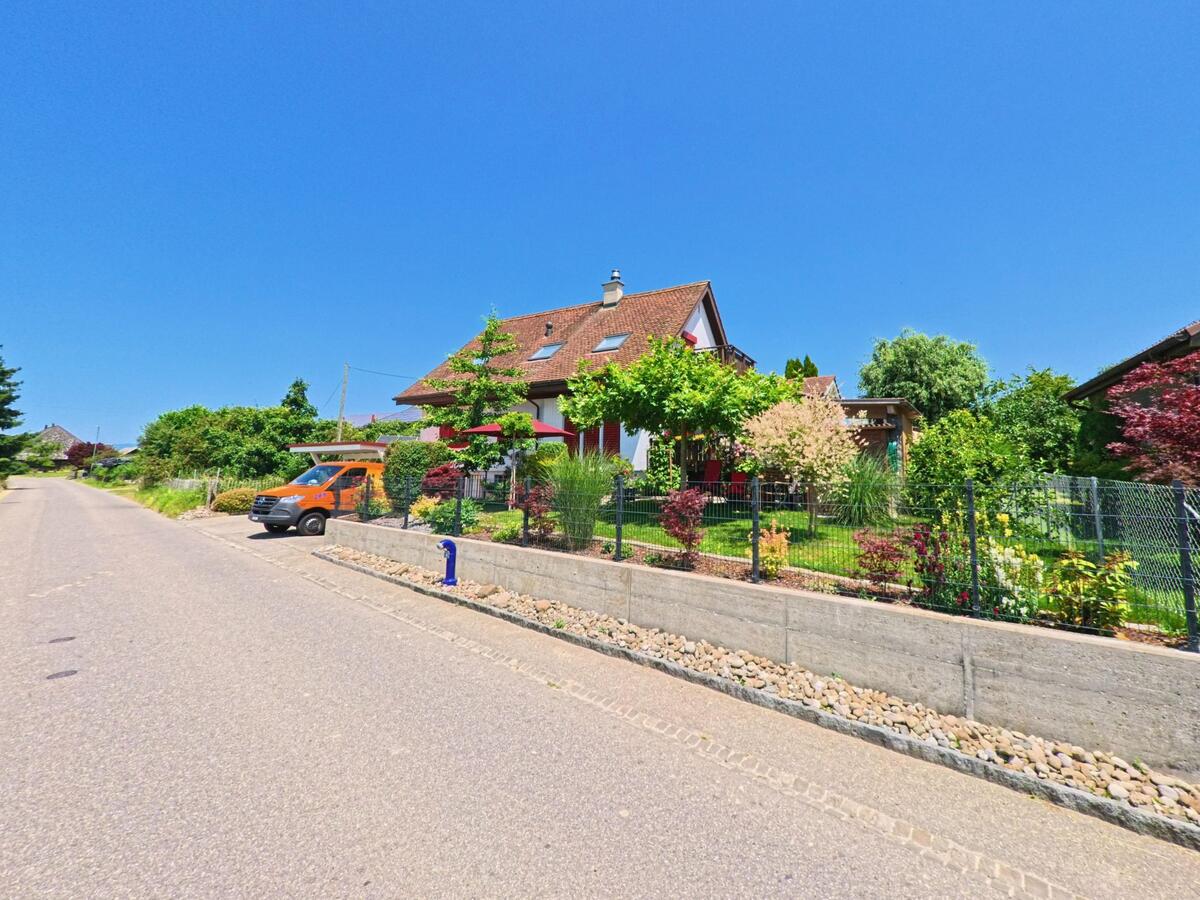 Ein modernes Haus in Iffwil (3305, Kanton Bern) mit einem gepflegten Garten und einem orangefarbenen Fahrzeug am Straßenrand.