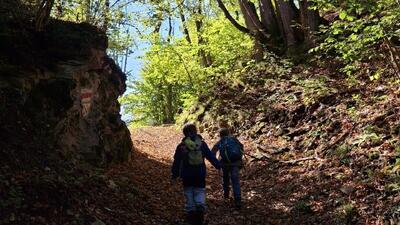 Zwei Kinder wandern auf einem mit Laub bedeckten Waldweg in Sarn (7423, Kanton Graubuenden) und folgen dem markierten Pfad des Erlebnsiwegs „Tilla“ durch das herbstliche Licht.