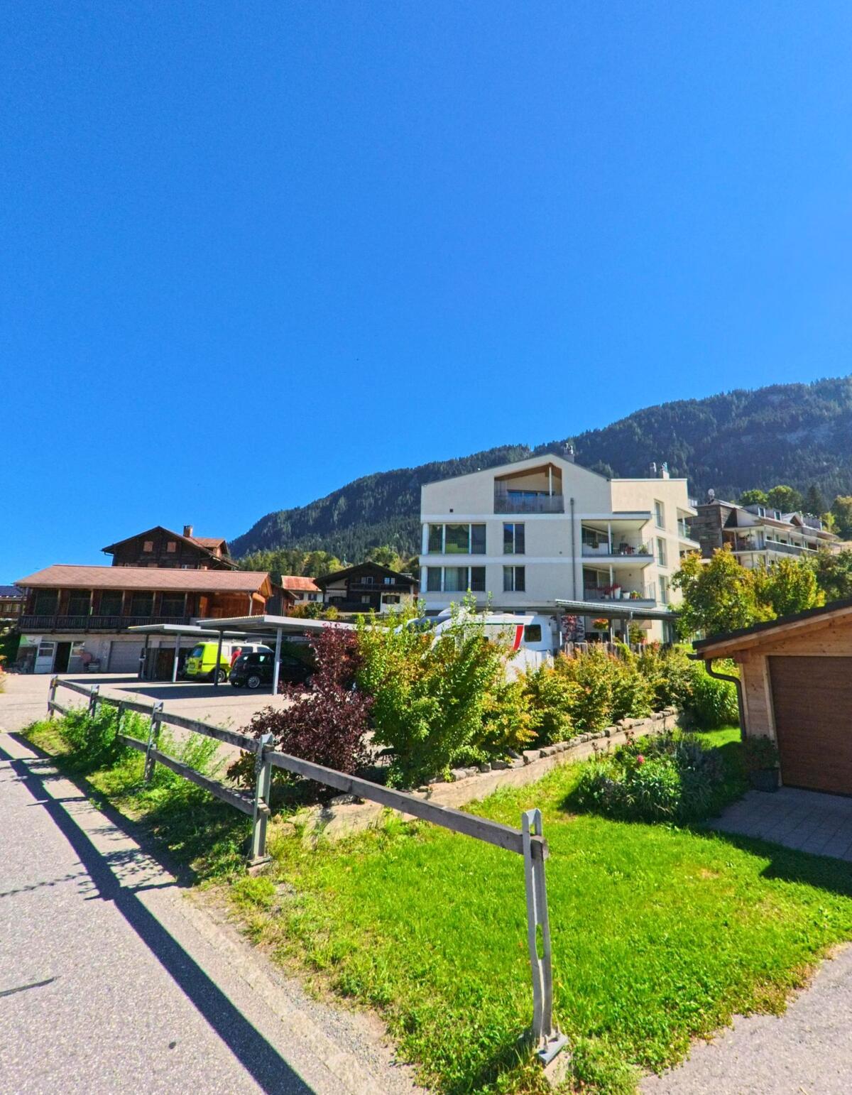 In Ilanz (7130, Kanton Graubünden) steht ein modernes Wohnhaus mit Balkonen am Hang, umgeben von traditionellen Holzhäusern und grüner Vegetation.