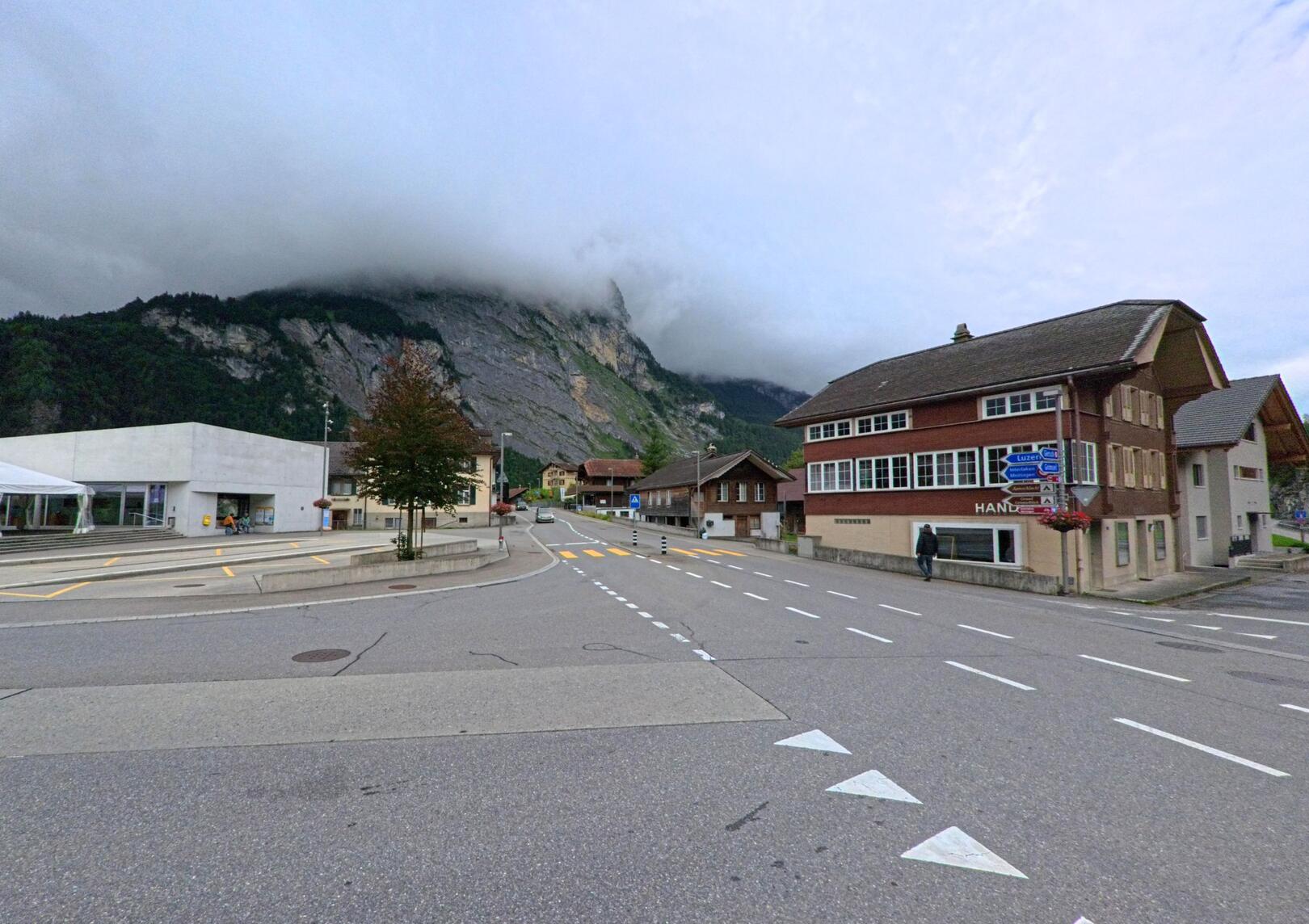 Im Zentrum von Innertkirchen (3862, Kanton Bern) treffen moderne und traditionelle Gebäude an einer breiten Strassenkreuzung aufeinander, vor einer eindrucksvollen, wolkenverhangenen Felswand.
