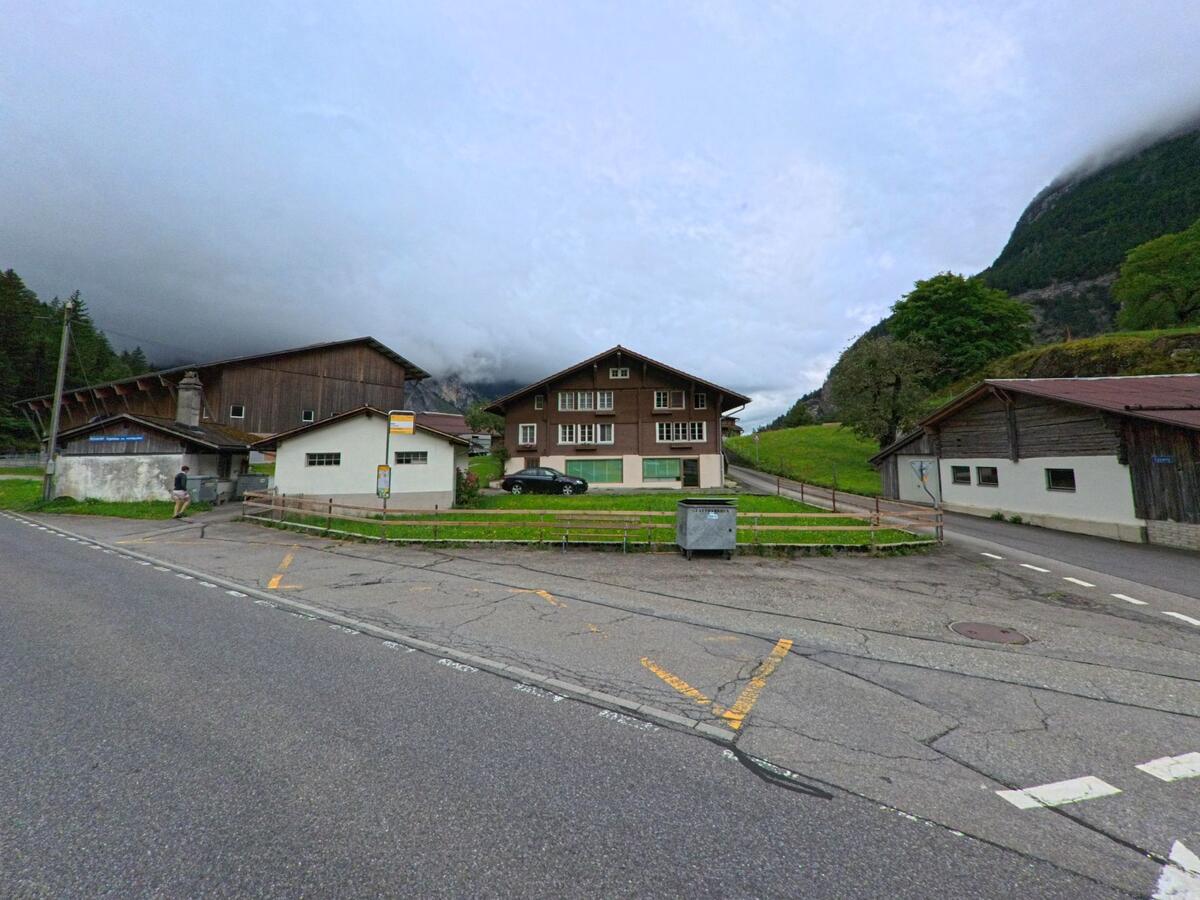 In Innertkirchen (3862, Kanton Bern) liegt eine Posthaltestelle an einer Kreuzung, umgeben von Bauernhäusern, Wiesen und bewaldeten Berghängen unter tief hängenden Wolken.