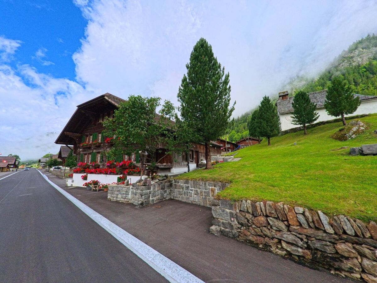 In Innertkirchen (3863, Kanton Bern) säumen ein traditionelles Bauernhaus mit roten Geranien und eine kleine Kapelle den Strassenverlauf am Hang vor nebelverhülltem Waldhang.