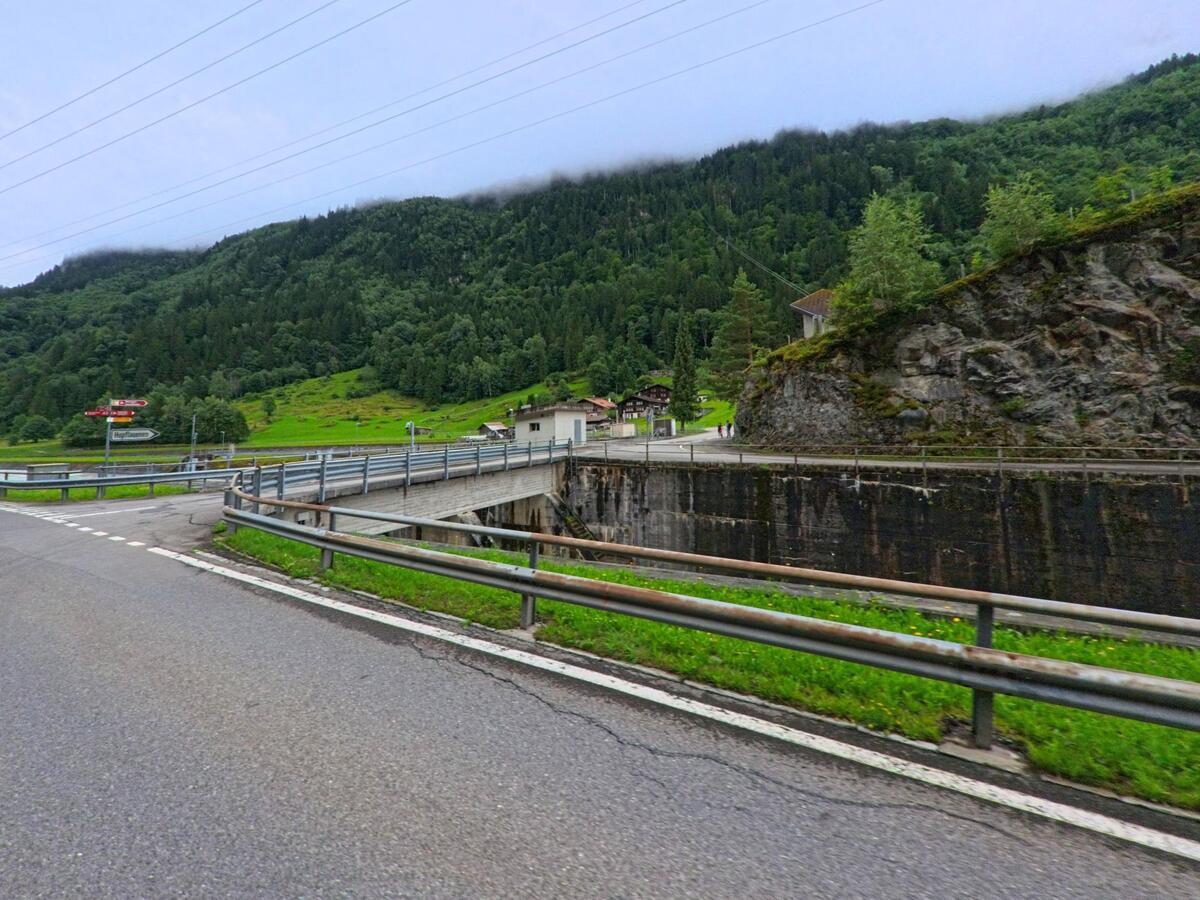 In Innertkirchen (3863, Kanton Bern) überspannt eine Brücke mit Leitplanken einen tief eingeschnittenen Kanal, eingerahmt von Felswänden und grüner Hanglandschaft mit verstreuten Gebäuden.
