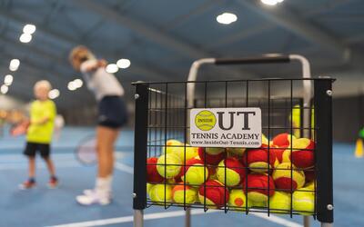 In Zürich (8092, Kanton Zürich) steht in einer Tennishalle ein Korb voller gelb-roter Tennisbälle im Vordergrund, waehrend im Hintergrund Kinder beim Training unscharf zu sehen sind und eine dynamische Sportsituation entsteht.