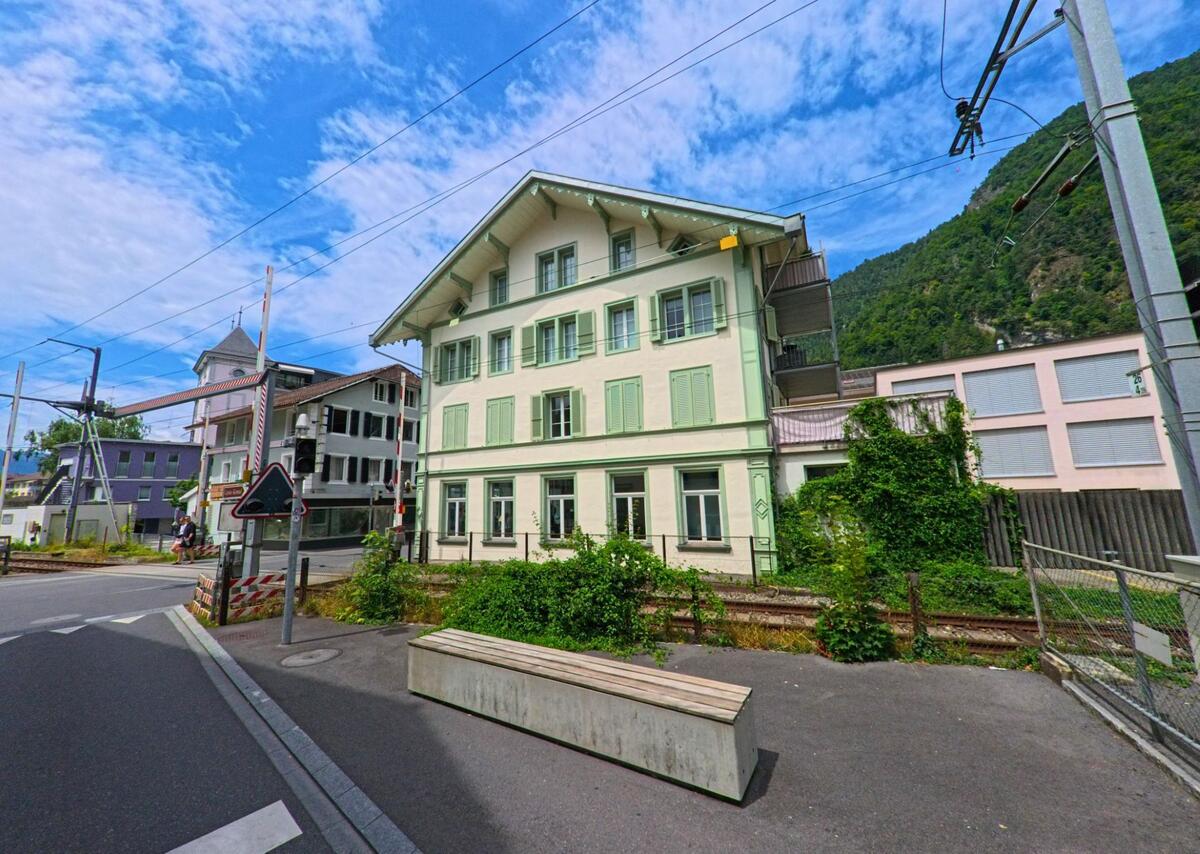 In Interlaken (3800, Kanton Bern) steht ein gruen-weiss gestrichenes Wohnhaus mit Fensterlaeden direkt an einer Bahnlinie und einer kleinen Sitzbank vor der Gleisanlage.