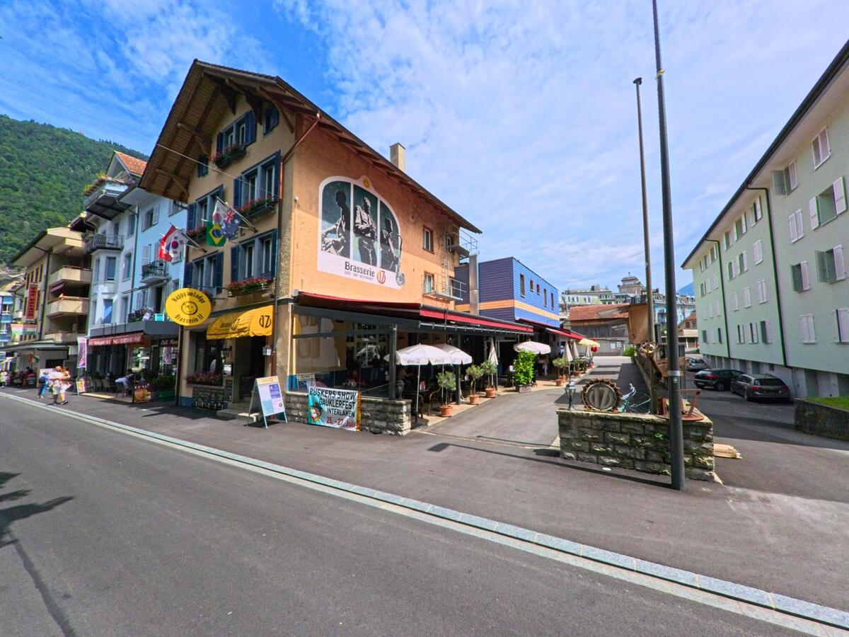 In Interlaken (3800, Kanton Bern) reihen sich entlang einer schmalen Strasse farbige Hauser mit internationalen Flaggen, Cafés und einer Brasserie mit Wandbild.