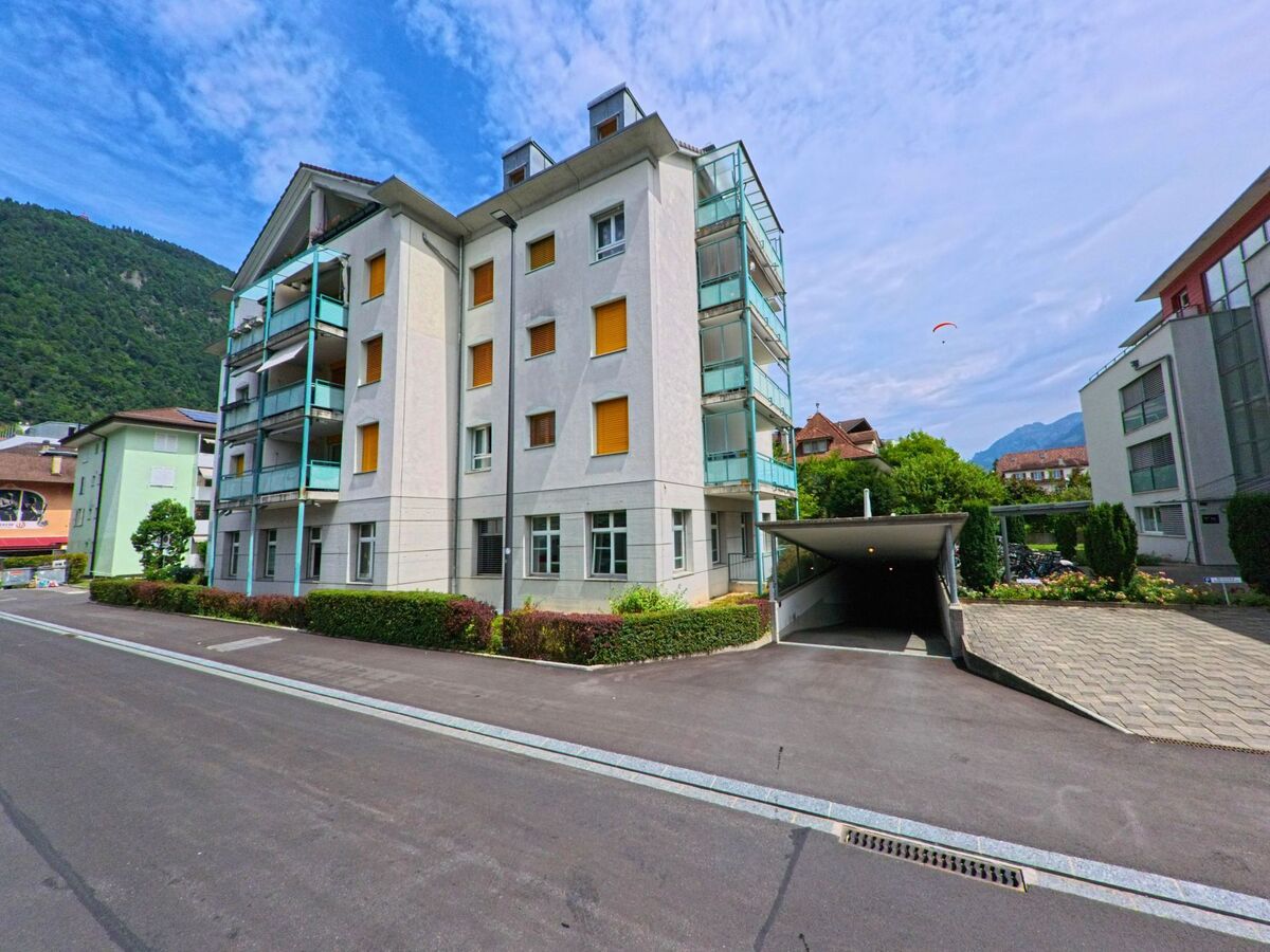 In Interlaken (3800, Kanton Bern) steht ein mehrgeschossiges Wohnhaus mit gelben Fensterlaeden an einer ruhigen Strasse, im Hintergrund erheben sich die Berge.