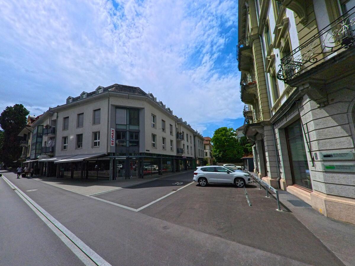 In Interlaken (3800, Kanton Bern) trifft eine moderne Wohn- und Geschaeftszeile mit dem Schriftzug „REMAX“ auf historische Fassaden entlang einer ruhigen Strasse mit Parkmoeglichkeiten.