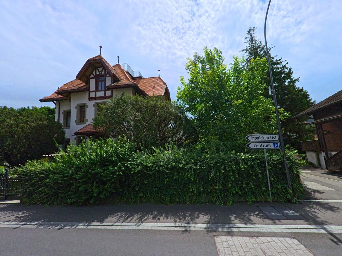 In Interlaken (3800, Kanton Bern) steht ein historisches Wohnhaus mit rotem Satteldach hinter dichter Vegetation, daneben zeigen Wegweiser die Richtung zum Zentrum und nach Interlaken Ost.