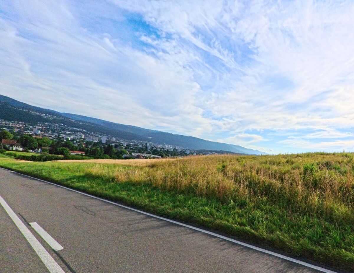 In Ipsach (2563, Kanton Bern) führt die Straße entlang einer Wiese mit weiten Ausblicken auf die umliegende Landschaft und den Himmel.