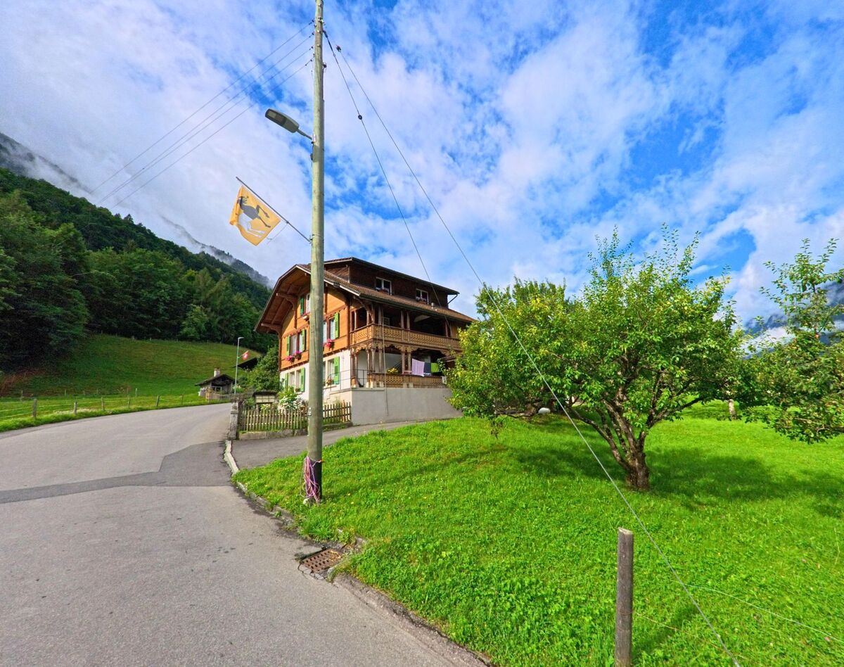 Am Rand einer Wiese mit Obstbäumen steht in Iseltwald (3807, Kanton Bern) ein traditionelles Holzhaus mit grün-roten Fensterläden unterhalb eines bewaldeten Hangs.