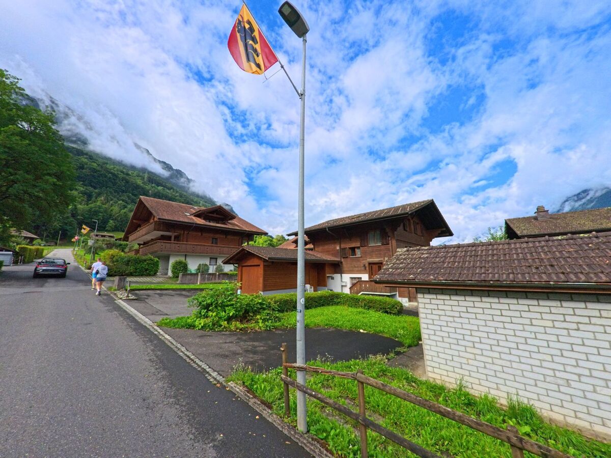 In einem ruhigen Quartier von Iseltwald (3807, Kanton Bern) führen mehrere Berner Fahnen entlang einer Wohnstrasse mit traditionellen Chalets am Fuss eines bewaldeten Hangs.