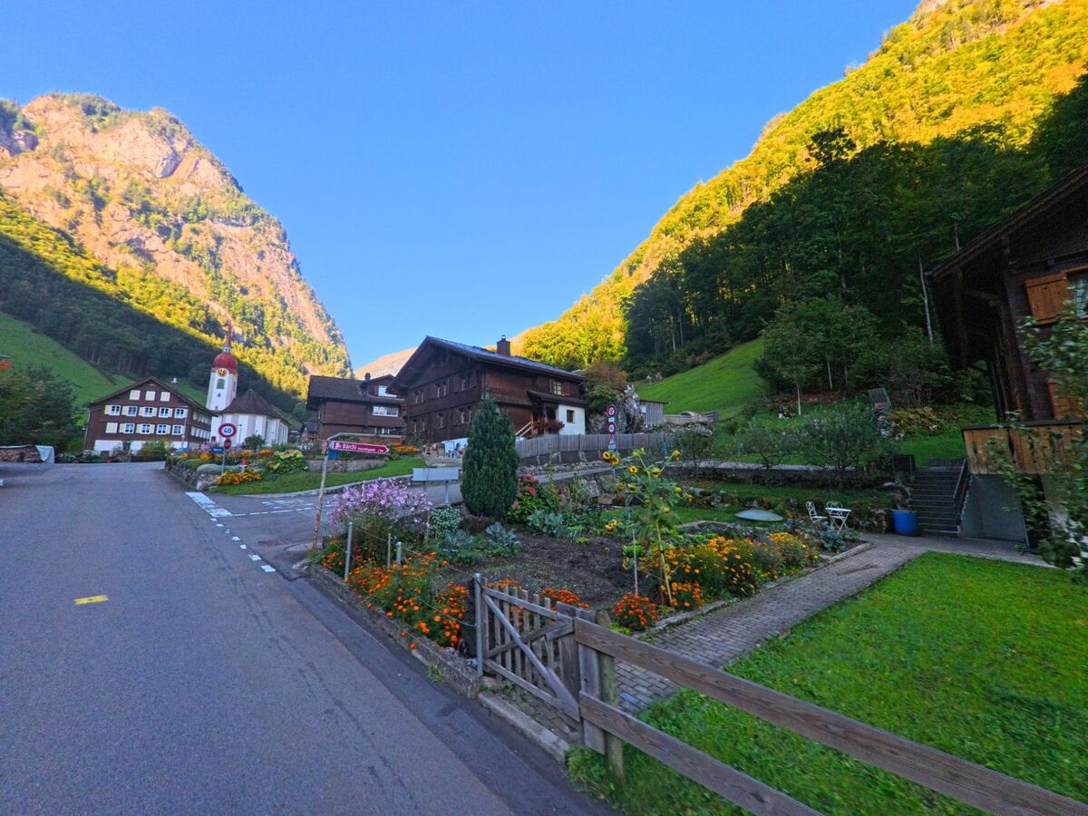 Entlang einer Dorfstrasse zeigt sich in Isenthal (6461, Kanton Uri) ein gepflegter Garten mit Blumenbeeten, umgeben von traditionellen Häusern und steilen Berghängen.