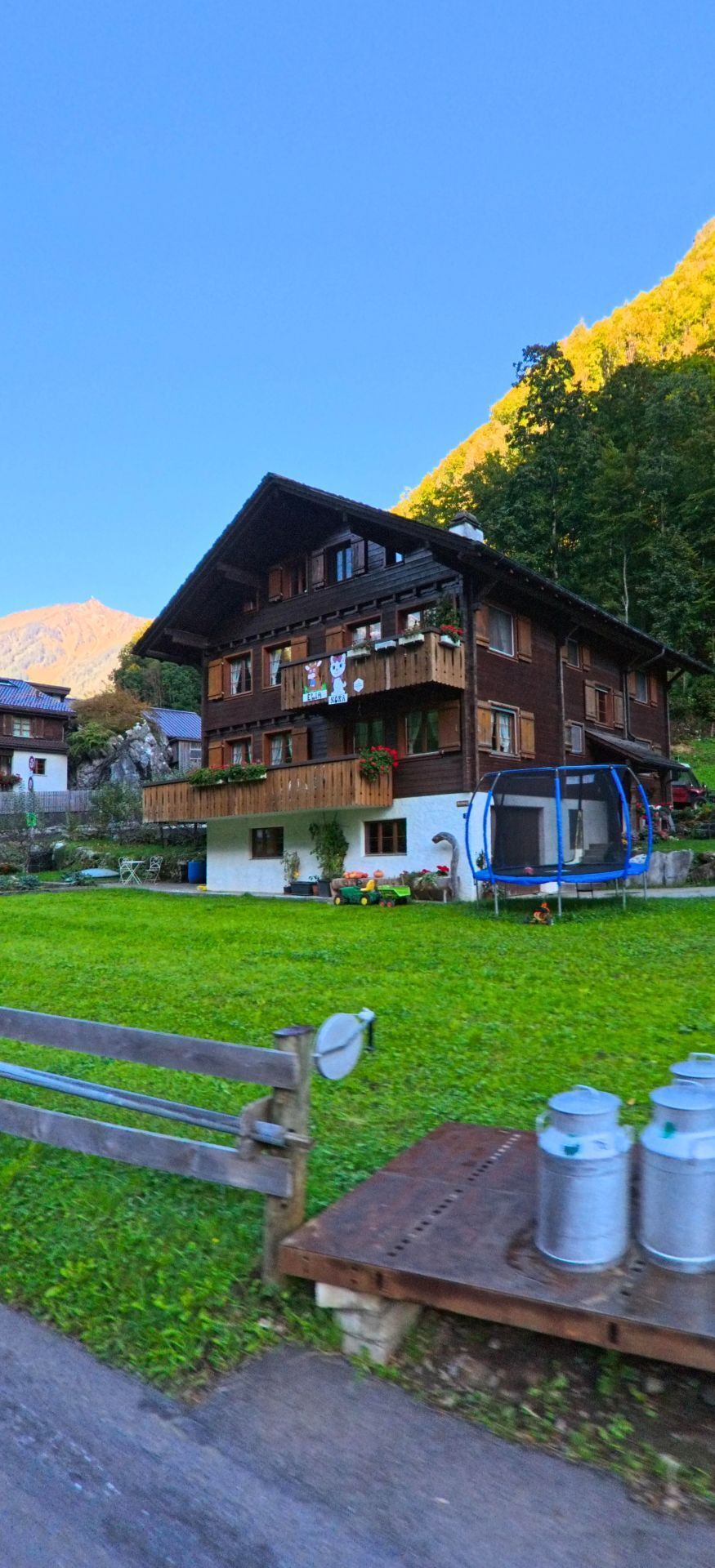Vor einem gepflegten Garten steht in Isenthal (6461, Kanton Uri) ein traditionelles Holzwohnhaus mit Balkon, Spielgeräten und angrenzender Wiese am Hang.