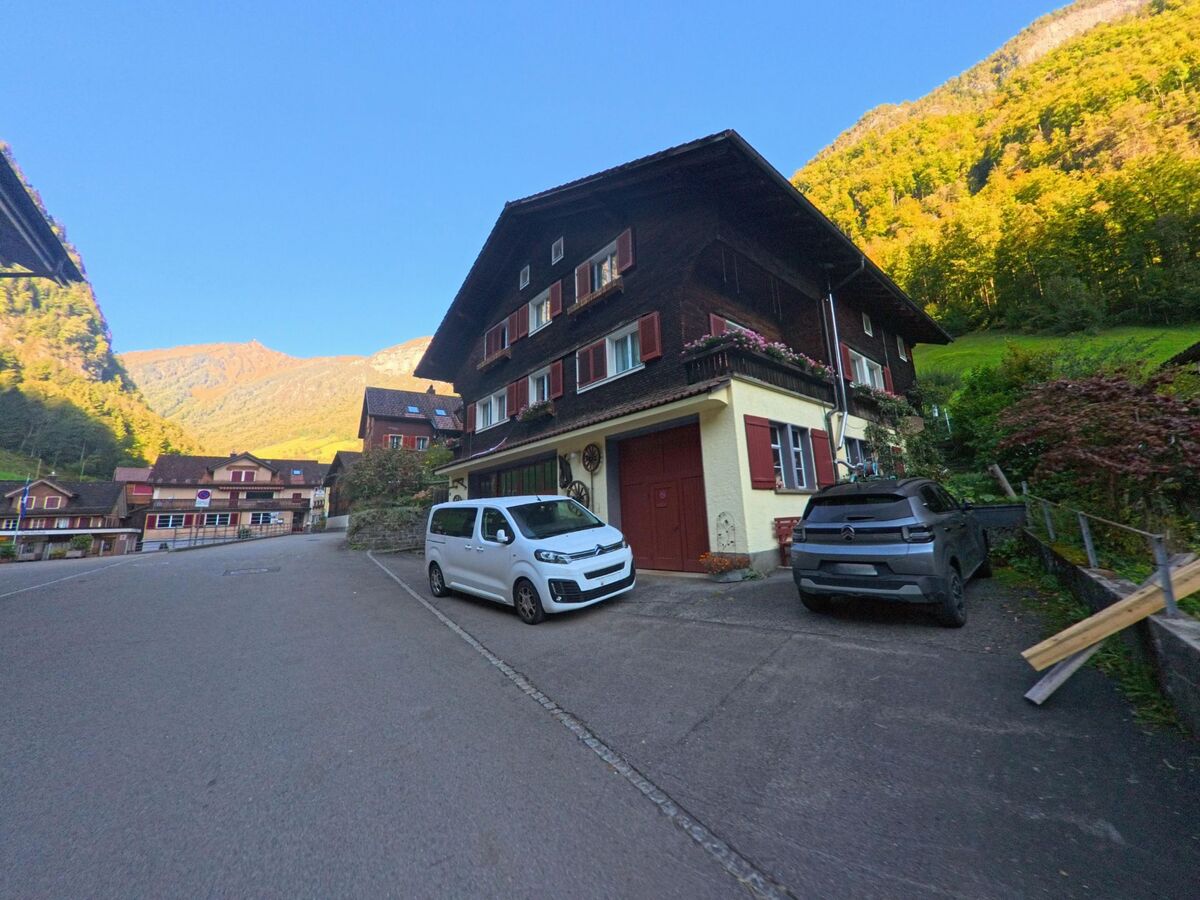Neben parkierenden Fahrzeugen steht in Isenthal (6461, Kanton Uri) ein traditionelles Mehrfamilienhaus mit Holzfassade an einer ruhigen Dorfstrasse vor bewaldeten Berghängen.