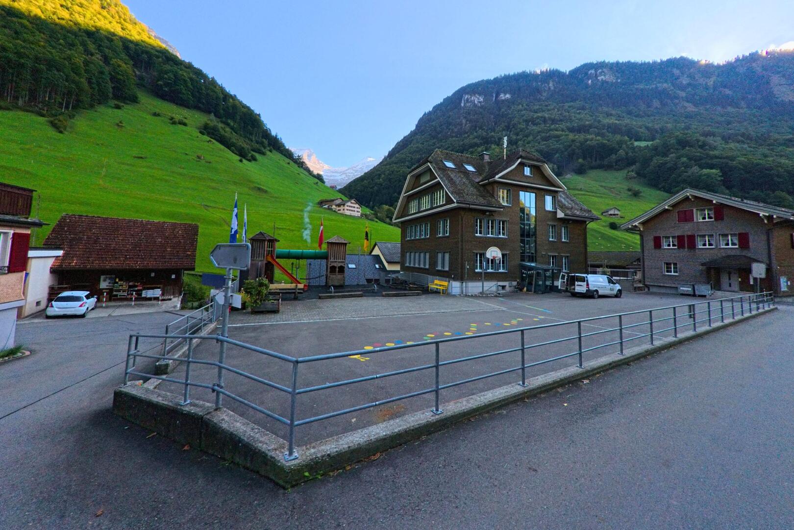 Vor grünen Berghängen liegt in Isenthal (6461, Kanton Uri) ein Schulareal mit Pausenplatz, Spielgeräten und mehreren Gebäuden im Talboden.