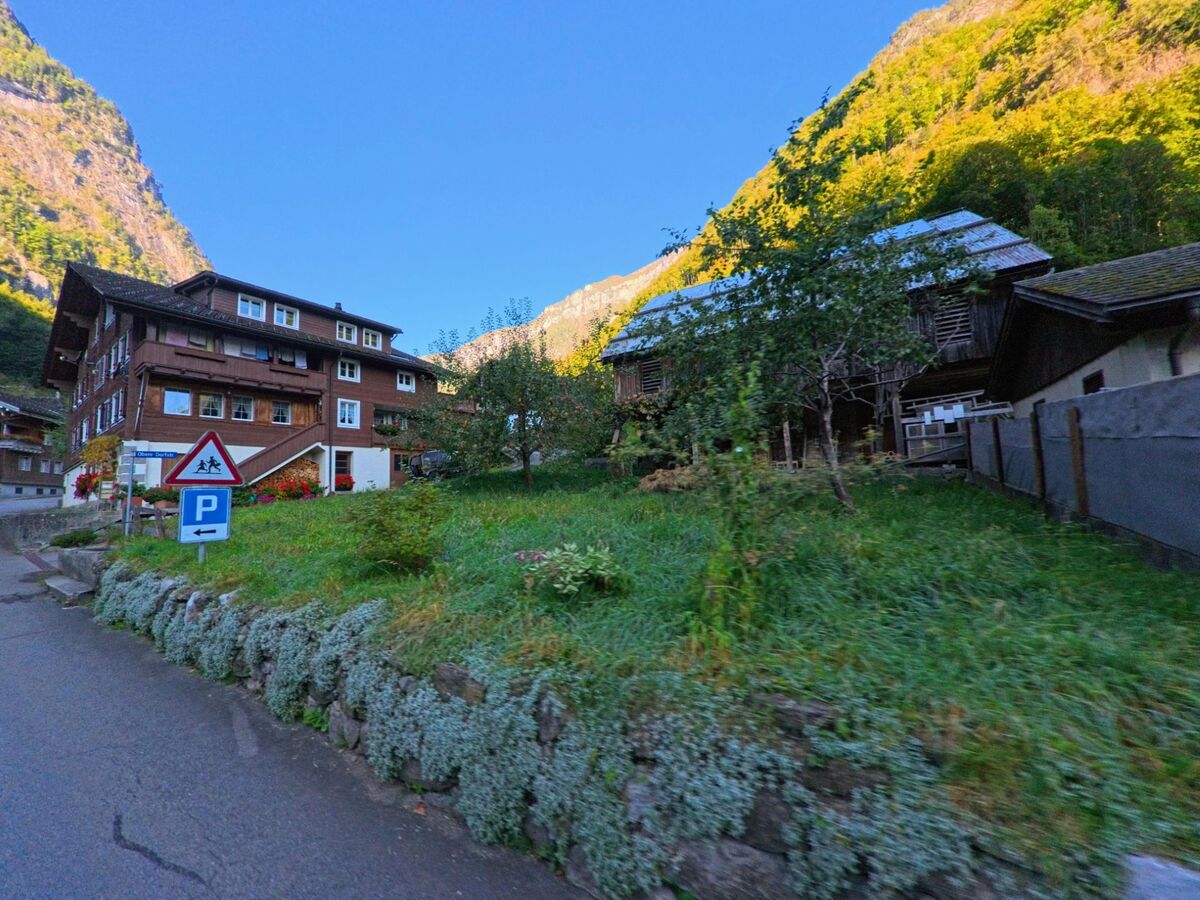 Am Strassenrand zeigt sich in Isenthal (6461, Kanton Uri) eine Wohnsituation mit traditionellem Mehrfamilienhaus, Wiese und landwirtschaftlichen Nebengebäuden vor steilen Berghängen.