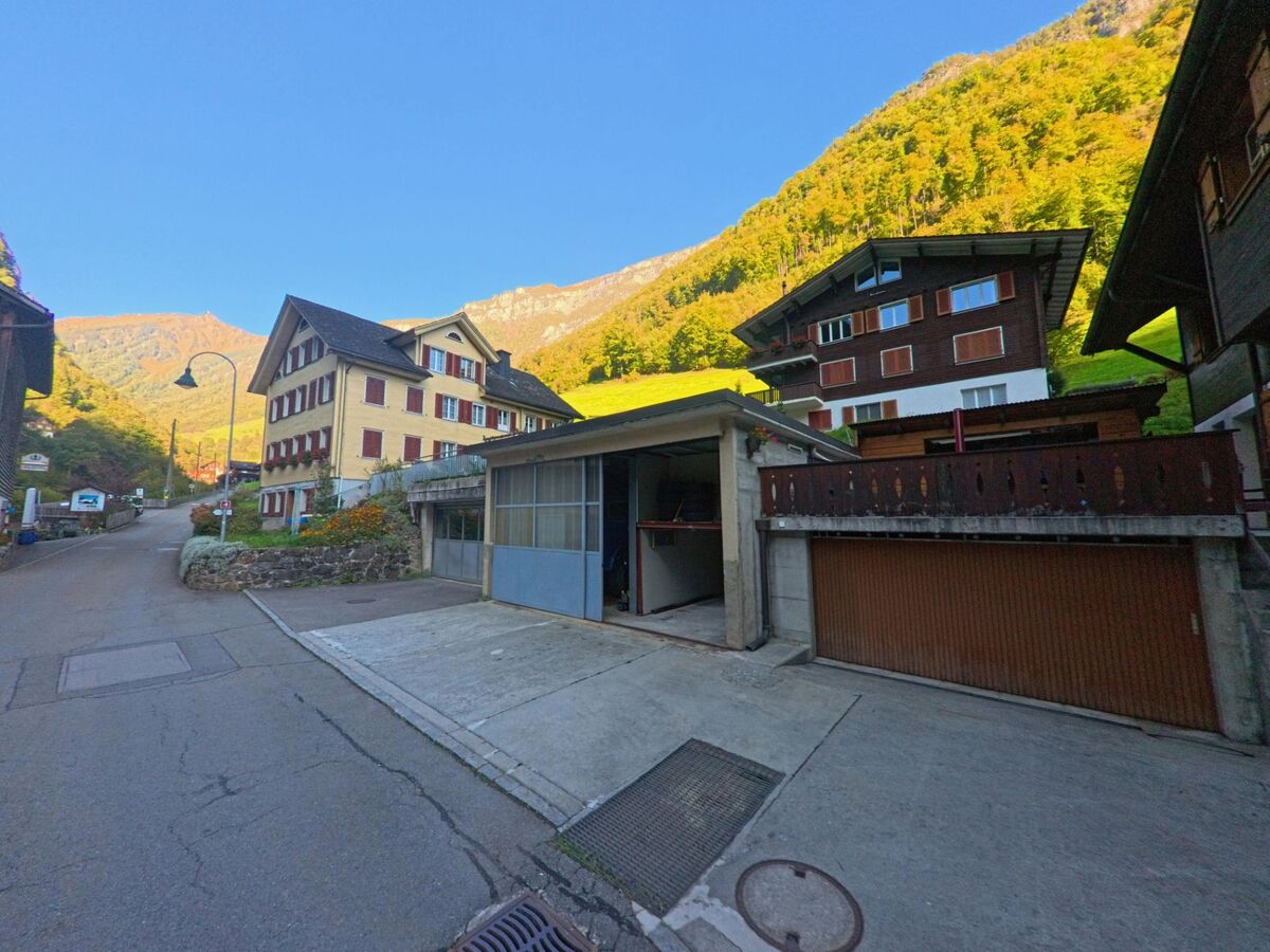 Entlang einer ansteigenden Strasse sind in Isenthal (6461, Kanton Uri) Wohnhäuser mit Garagen und Blick auf sonnige Berghänge zu sehen.